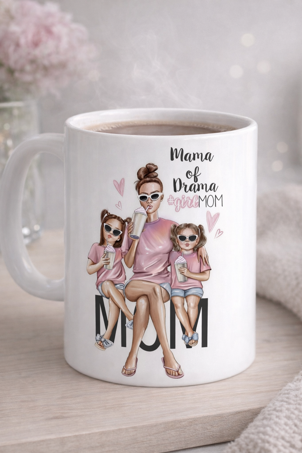 Ceramic white coffee mug featuring a stylish illustrated mom sitting with her two kids, wearing sunglasses, with the text “Mama of Drama” printed on the front.
