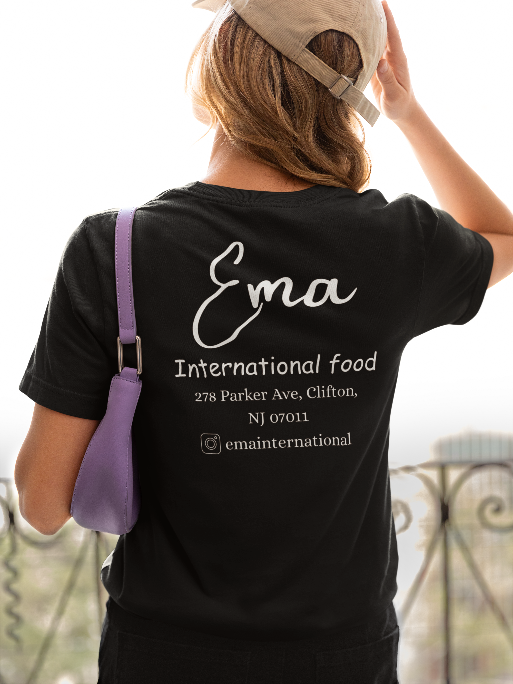 Back view of a ManiaWear custom T-shirt worn by a woman standing near a balcony, showcasing the shirt's fit and design.